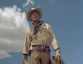 A tall, lean white man in his fifties dressed in Western clothes, mostly brown, with a dark blue neckerchief and a brown cowboy hat. He's wearing a gunbelt slung low on his hips and stands against a blue sky with one large white cloud.