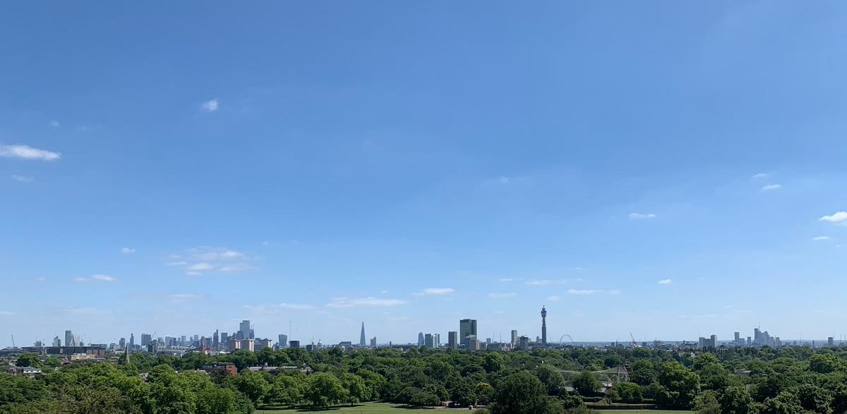 A photography of the skyline of London, seen very low and far away from Primrose Hill in the north. Above is a blue sky.