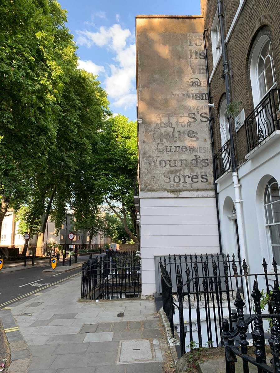 A photo of a building on a London street. The top two-thirds of it are brick and show the faded remains of an advertising sign. Most of the words are illegible, but towards the bottom you can make out "Cures" and "Wounds and Sores."